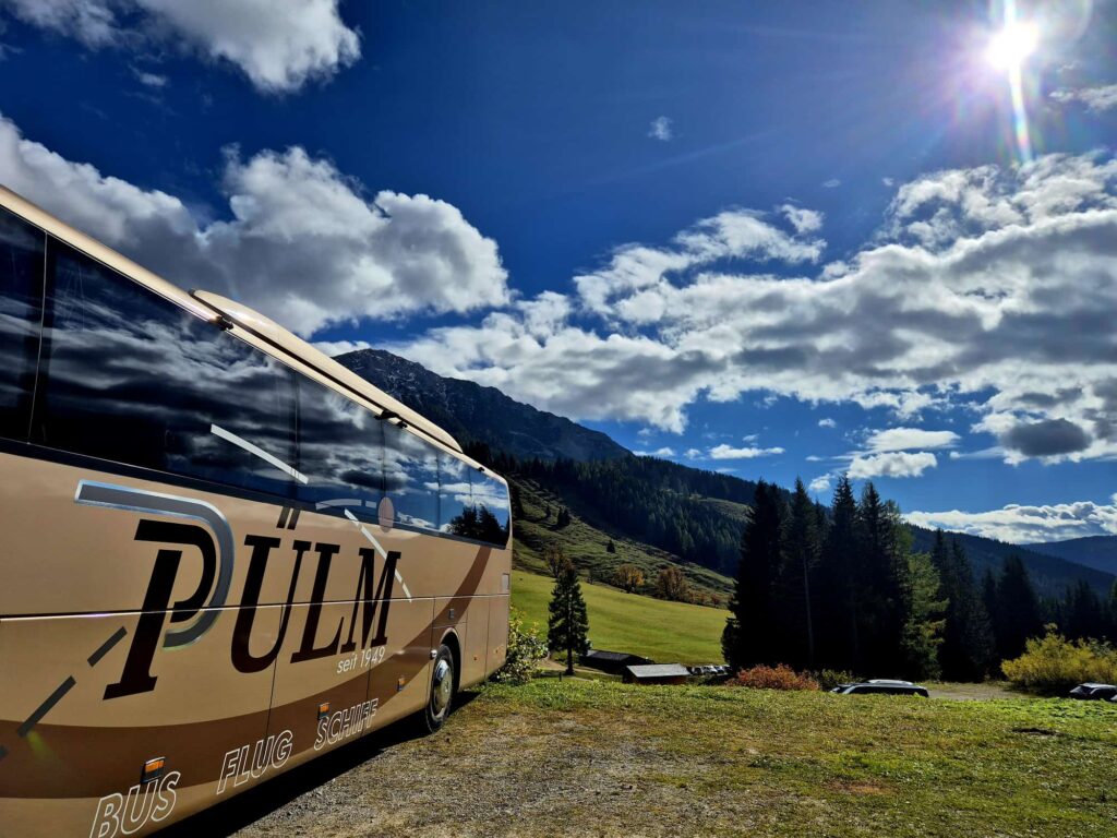Ein Reisebus der Firma Pülmreisen vor Bergpanorama mit Tannenwäldern.