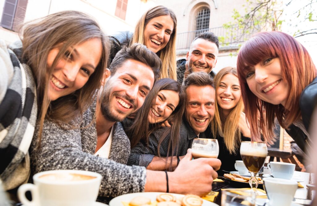 Eine Gruppe junger Menschen in einem Outdoor-Cafe lacht in die Kamera. Im Vordergrund stehen Kaffee und Kekse.