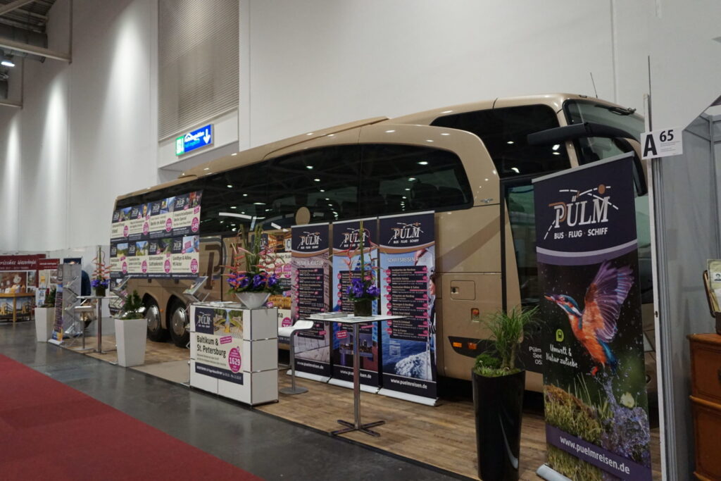 Ein Reisebus der Firma Pülmreisen auf einem Messestand mit Plakaten von Reiseangeboten.