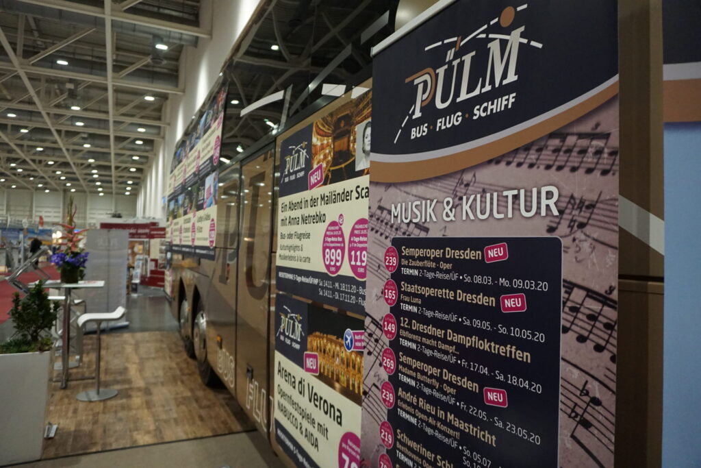 Ein Reisebus der Firma Pülmreisen auf einem Messestand mit Plakaten von Reiseangeboten.