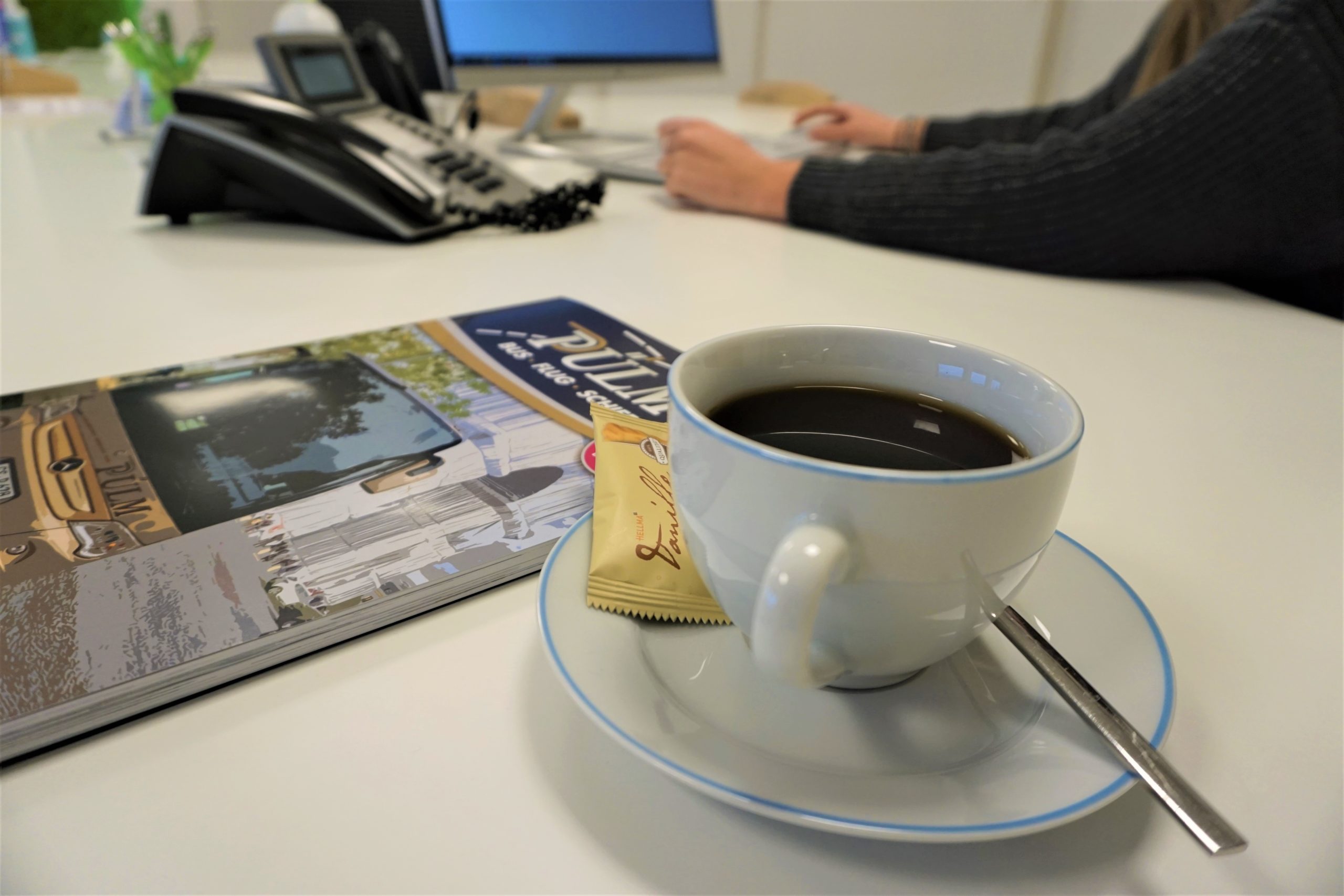 Eine Kaffeetasse und ein Katalog der Firma Pülmreisen auf dem Schreibtisch einer Mitarbeiterin.