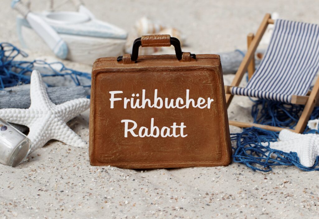 Ein brauner Koffer mit der Aufschrift Frühbucher Rabatt auf Sand vor einem Liegestuhl.