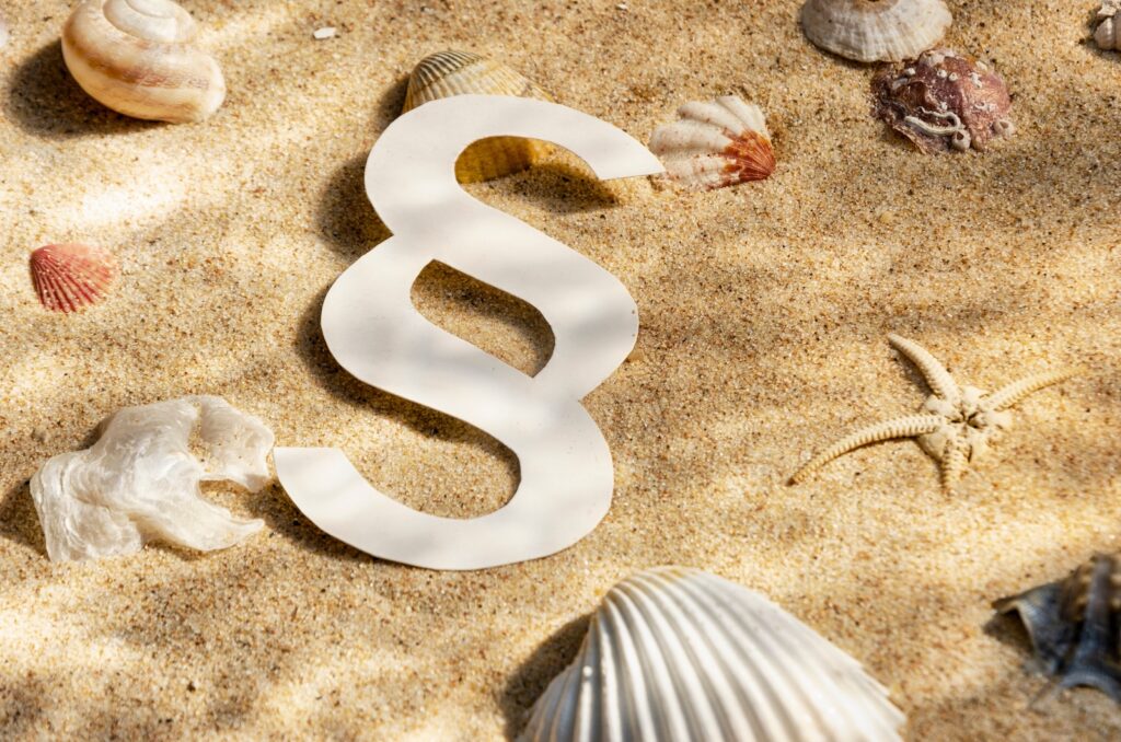 Ein Paragraphenzeichen auf Sand mit Muscheln.