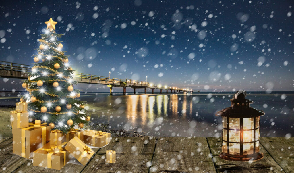Ein beleuchteter Weihnachtsbaum mit Geschenken im Schneetreiben vor einer Seebrücke am Meer.