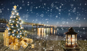 Ein beleuchteter Weihnachtsbaum mit Geschenken im Schneetreiben vor einer Seebrücke am Meer.