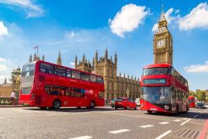 Zwei rote Busses fahren durch London vor Big Ben und Westminster Abbey.