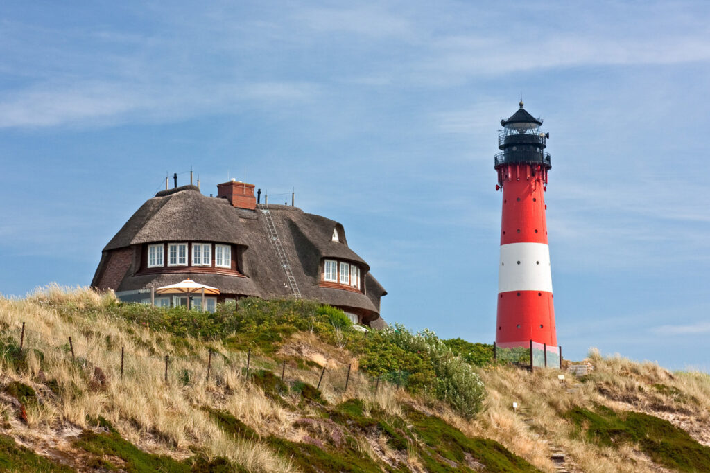 Rot-Weißer Leuchtturm in Hörnum auf der Insel Sylt an der Nordsee mit einem Friesenhaus im Hintergrund.
