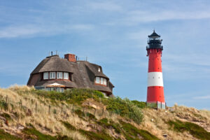 Rot-Weißer Leuchtturm in Hörnum auf der Insel Sylt an der Nordsee mit einem Friesenhaus im Hintergrund.